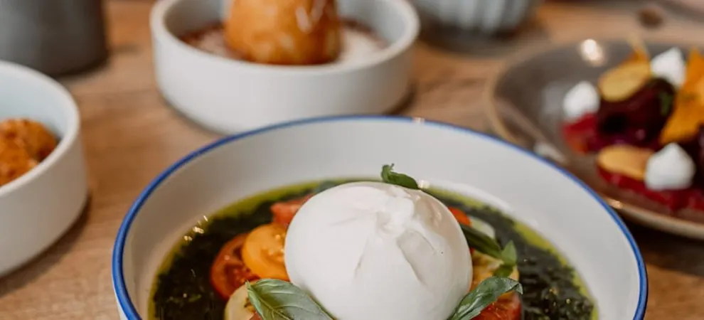 A bowl of soup with a poached egg, vegetables and herbs, surrounded by other dishes on a wooden table.