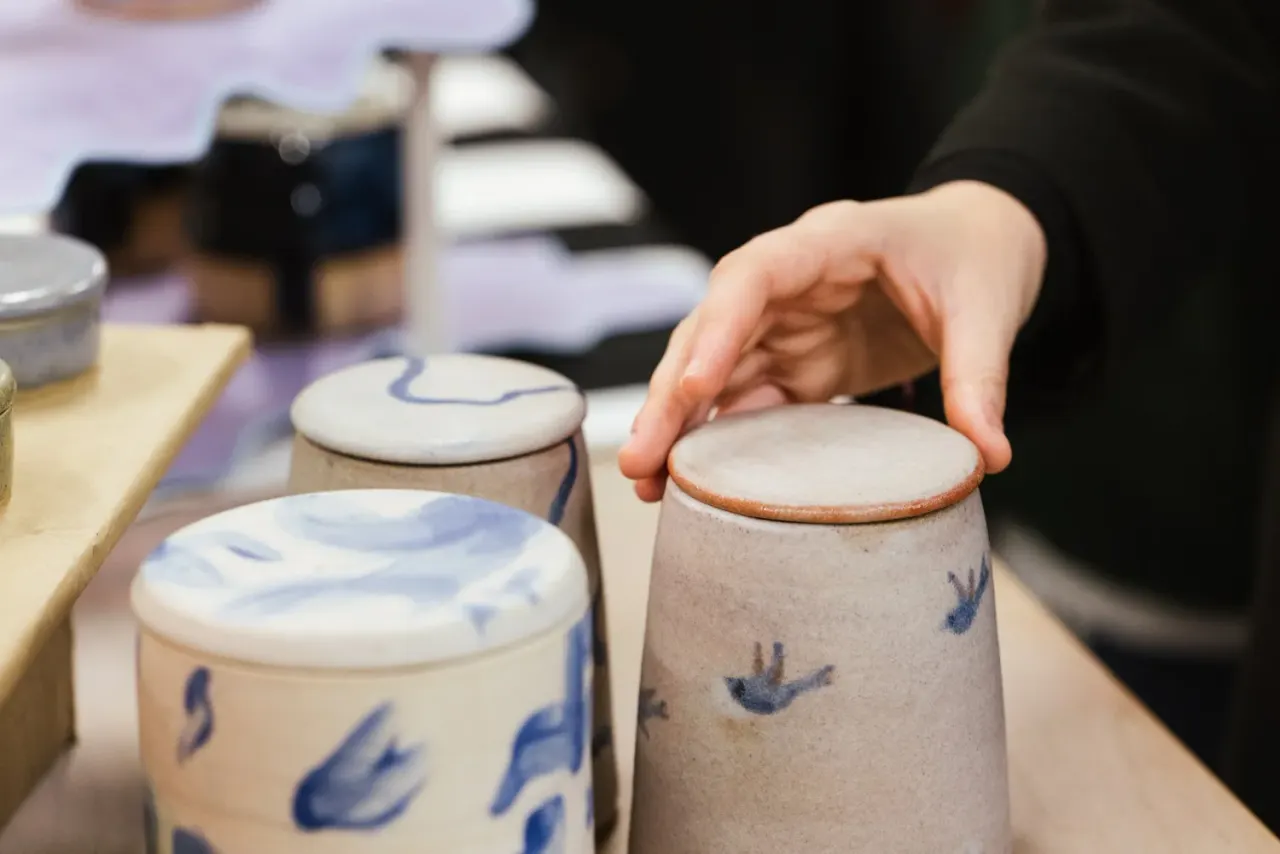 Person's hand touching a ceramic pot with blue decoration among other handmade pottery pieces on display.
