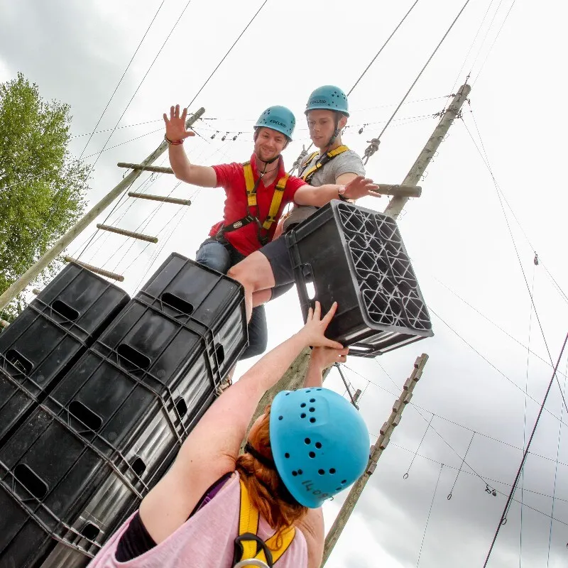 Two people in helmets and safety harnesses navigate a high ropes course obstacle above another participant below.