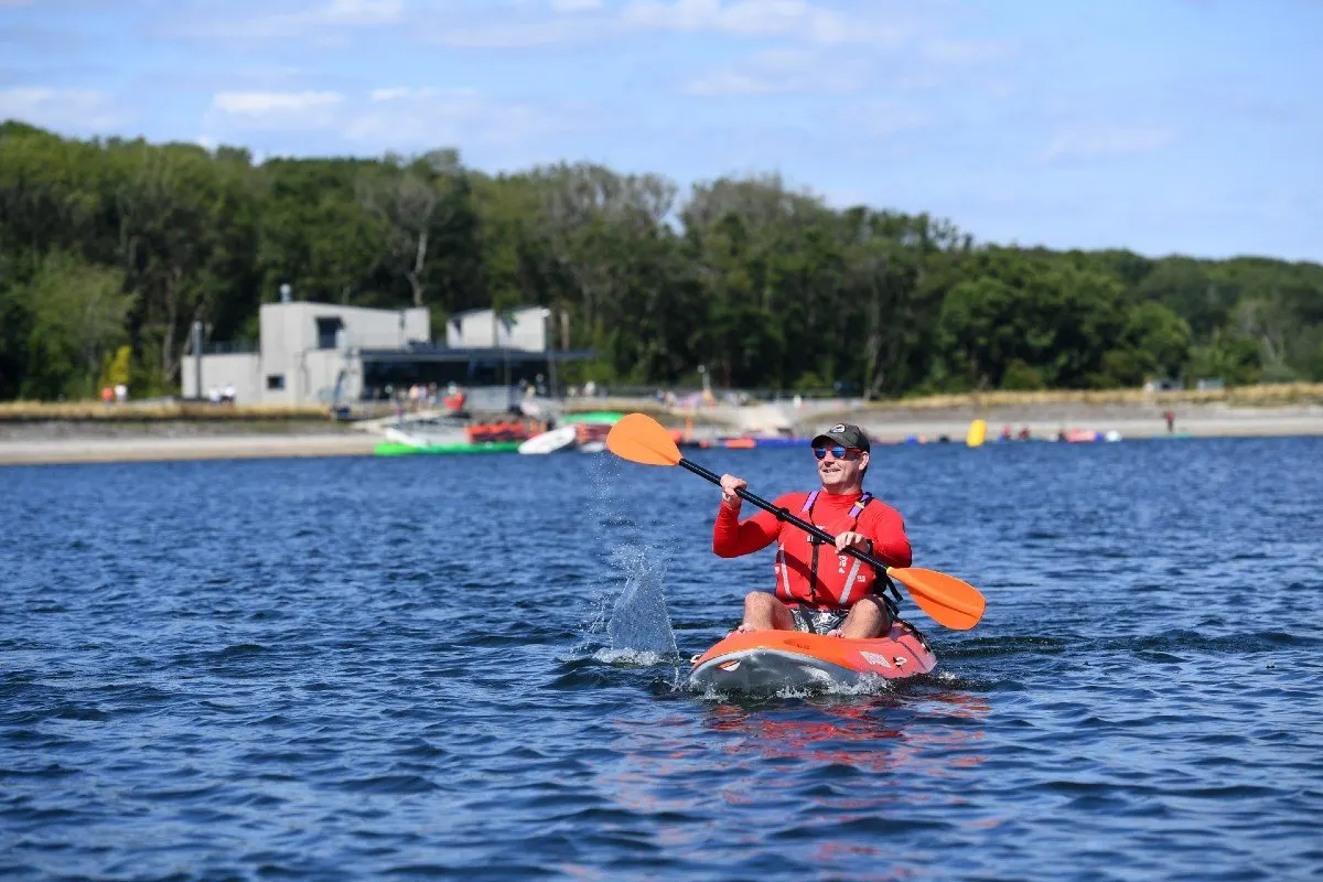 Man in red life jacket paddling orange kayak on reservoir with waterside building and trees behind.