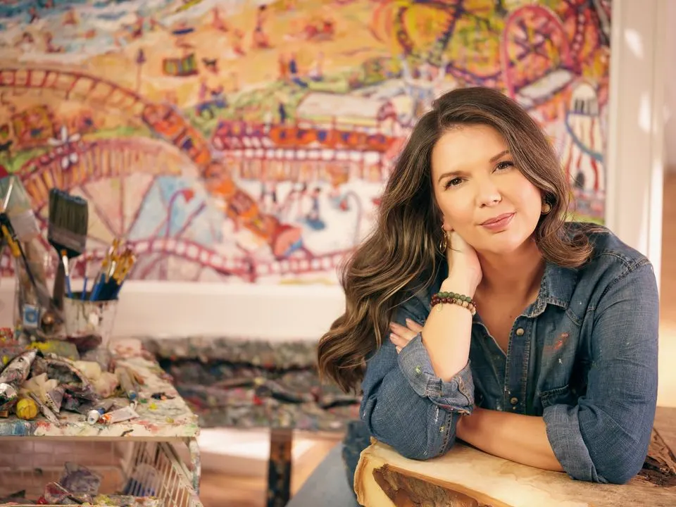 Woman posing in artist studio with vibrant fairground-themed painting in background.