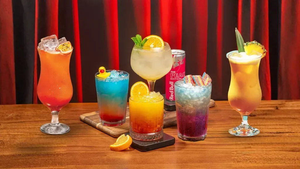 Colourful cocktails with ice, citrus slices and garnishes arranged on a wooden bar against red curtains.
