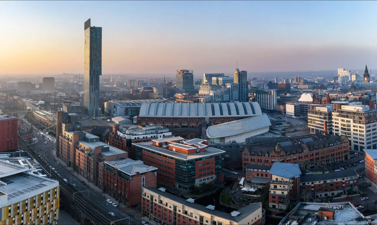 Aerial cityscape of Manchester at sunrise, showing the indoor arena and surrounding Victorian and modern buildings against a hazy skyline.