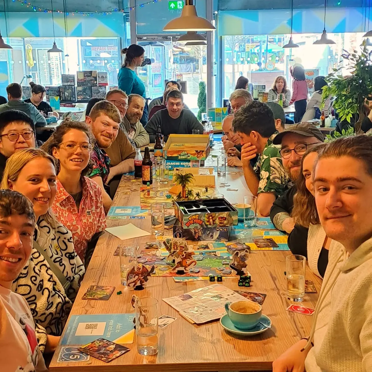 A group of adults and children gathered around long wooden tables playing board games whilst having drinks and food in a bright café space.