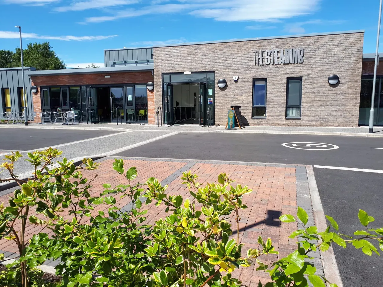 Modern brick leisure facility with car park, outdoor seating area, and landscaping on a sunny day.