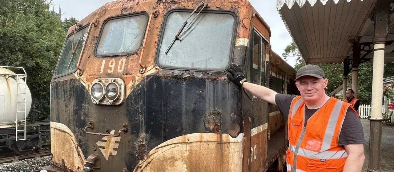 A man in an orange safety vest stands beside a weathered heritage railway diesel locomotive numbered 190 at a heritage railway depot.