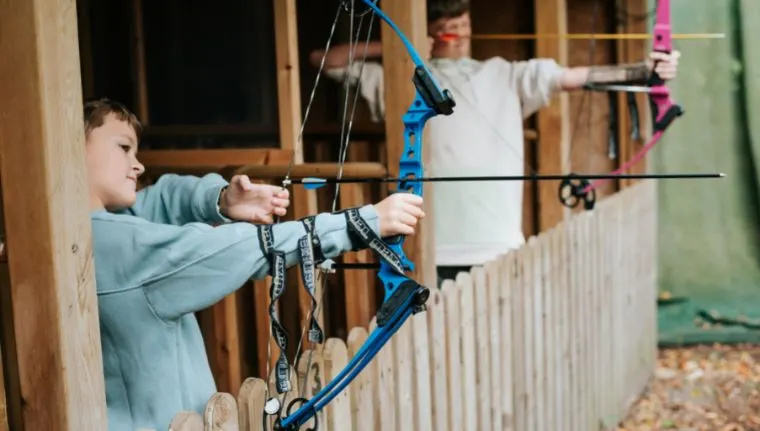 Young person drawing back a blue compound bow during an archery activity session indoors.