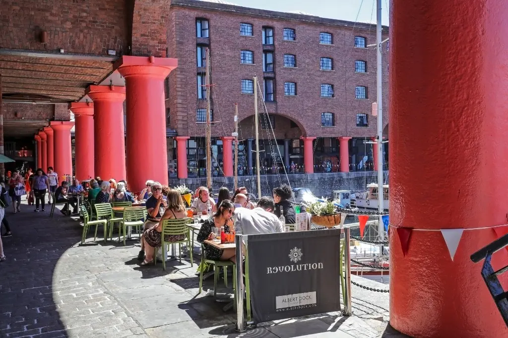 Outdoor seating area with green chairs and diners at a waterside venue surrounded by large red pillars and Victorian brick buildings.