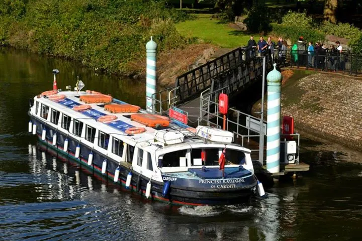 A passenger river boat moored at a pontoon with visitors boarding via a bridge alongside grassy banks.