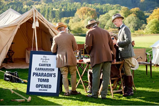 People in period costume stand near a canvas tent and historical display sign at an outdoor event beside a river and forested hillside.