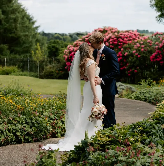 Bride and groom embrace on a garden path with flowering shrubs and lawn behind them.