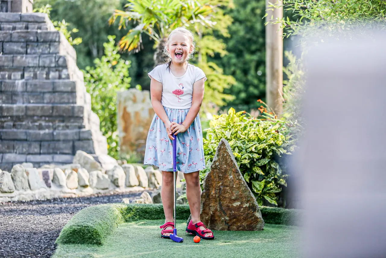 A young girl laughs while playing mini golf on an outdoor course surrounded by garden plants and stone features.