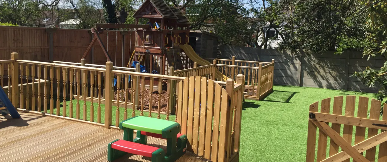 Outdoor children's play area with wooden climbing structures, slides, decking, artificial grass, picnic table and wooden fencing.