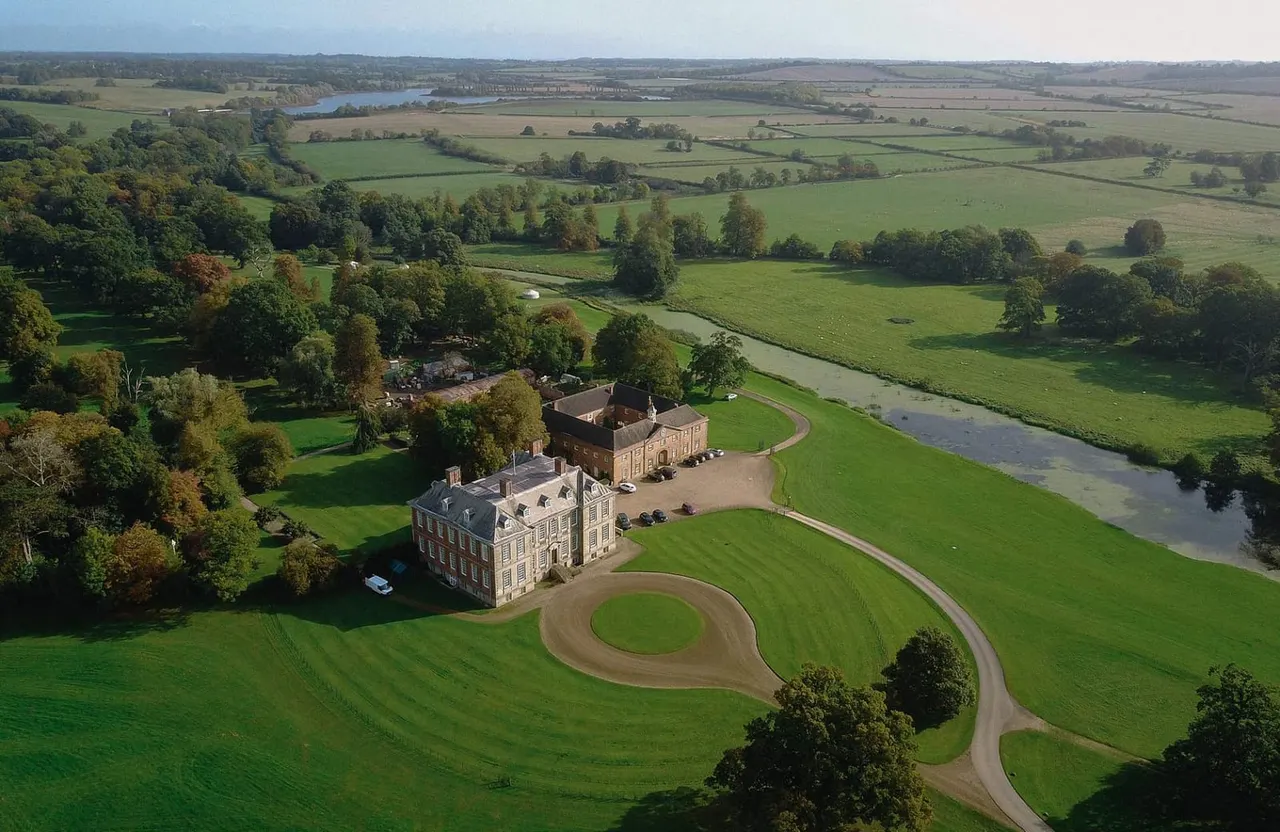 Aerial view of a grand country house with formal grounds, tree-lined estate and river, surrounded by rolling countryside and farmland.