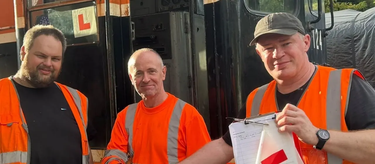 Three men in high-visibility orange clothing stand in front of a coach, one holding a clipboard.