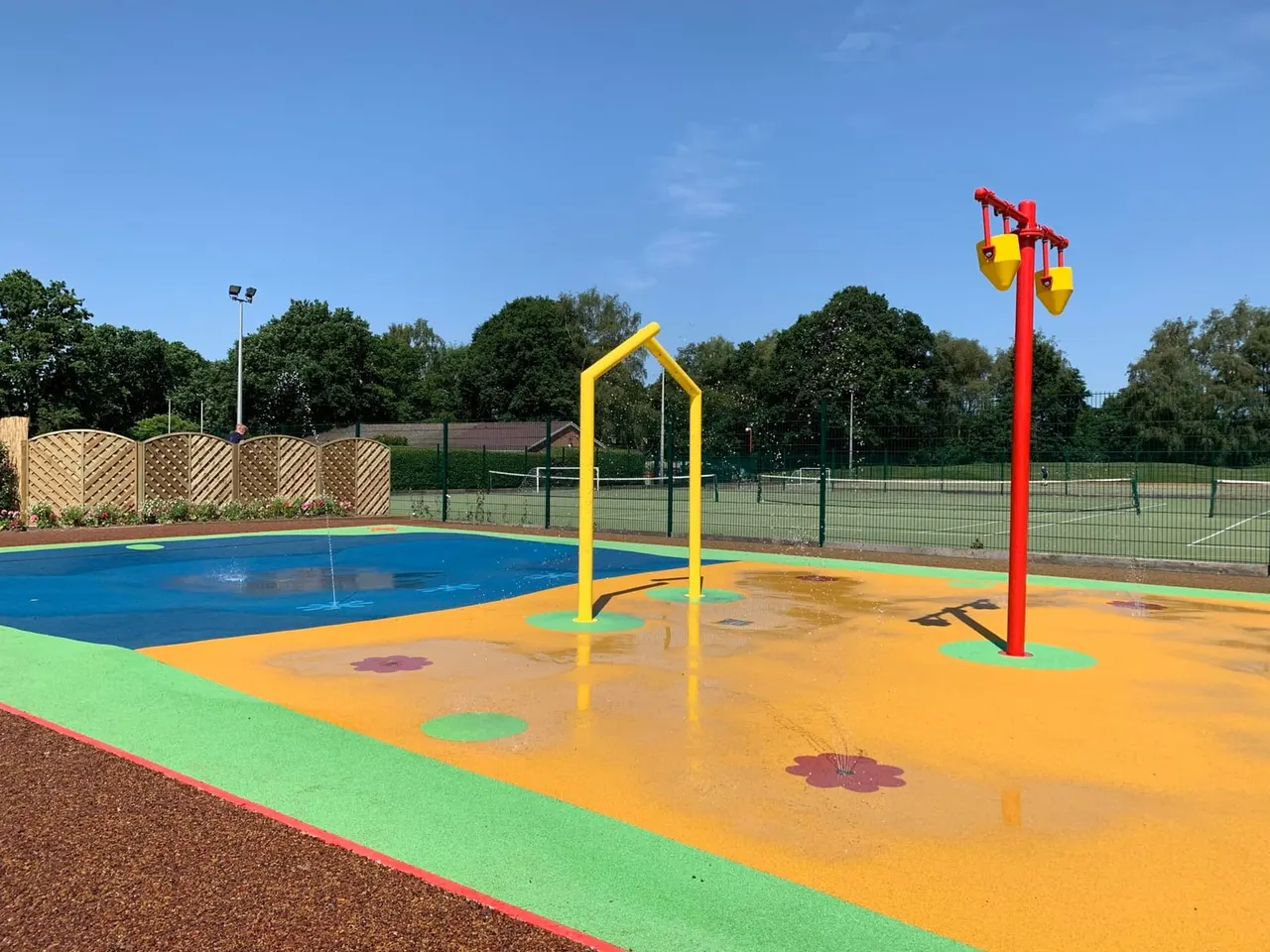 Colourful outdoor splash pad with yellow archway and red water play structures, surrounded by sports courts and trees.