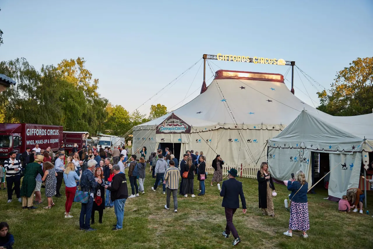 Crowds of visitors walk around a traditional circus big top and supporting tents on an autumn grass field.