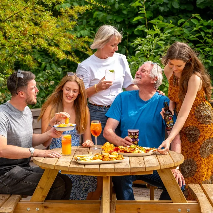 Five adults socialising around a wooden picnic table with food and drinks in a garden setting.