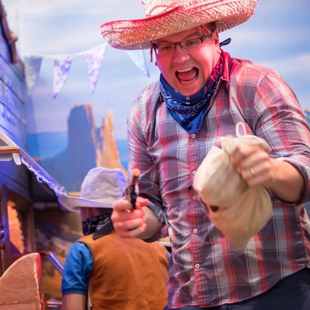 Man in sombrero and checked shirt entertains a young child indoors with Western-themed décor.