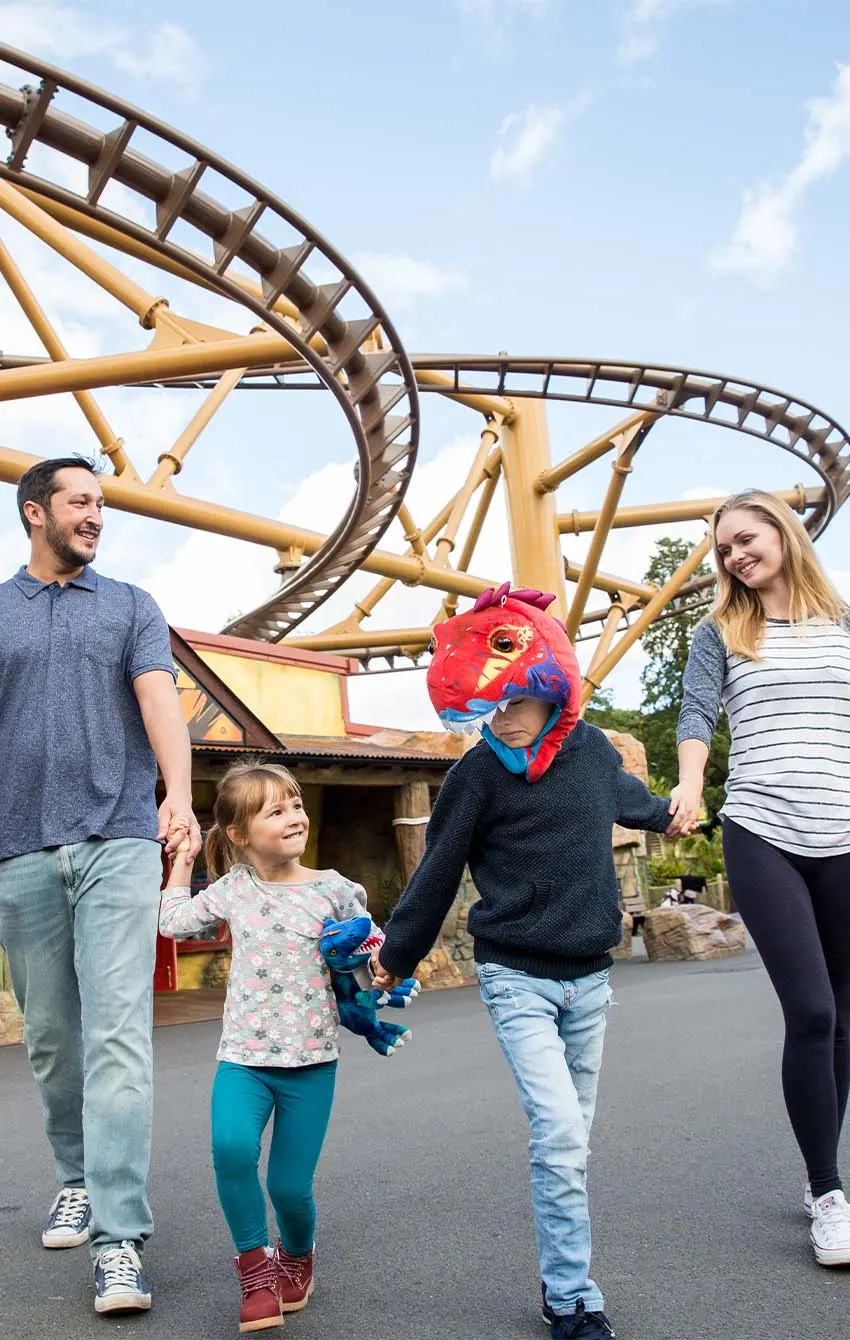 Family walking together in front of a large roller coaster ride with yellow support structures.