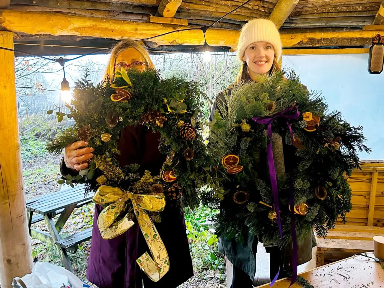 Two people holding large festive wreaths decorated with greenery, dried citrus slices and ribbons under a wooden shelter.