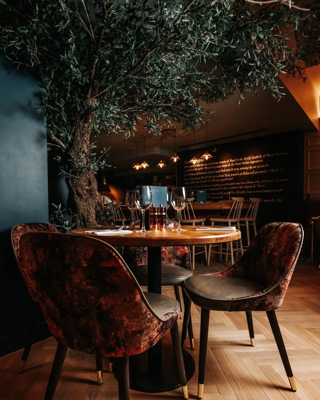 Upmarket restaurant dining area with a round wooden table, velvet burgundy chairs, wine glasses, and a large olive tree overhead under warm pendant lighting.