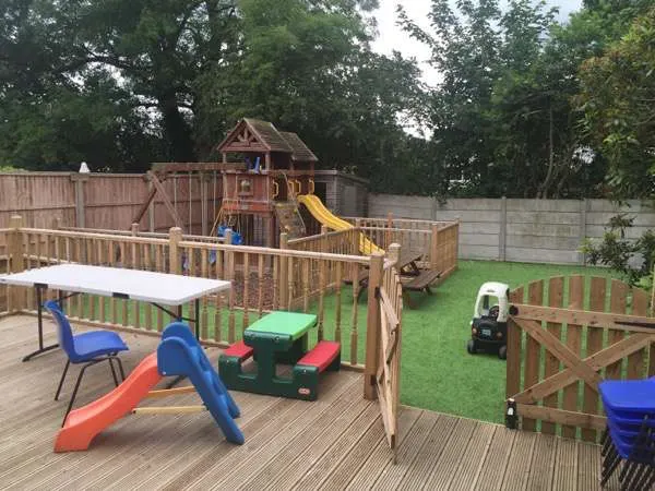 Outdoor play area with wooden decking, climbing structure, slides, and various play equipment for young children on grass surrounded by fencing.