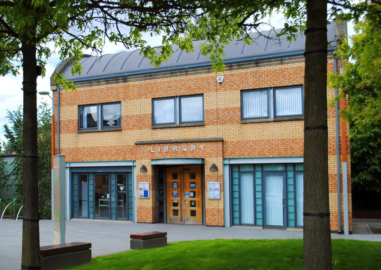 Red brick library building with blue trim and entrance doors, framed by tall trees with green foliage.