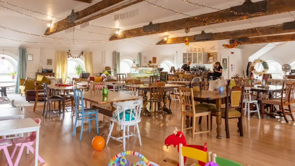 Spacious, bright café interior with wooden beams, string lights and bunting, multiple tables and chairs in various colours, children's toys scattered on wooden floor, and staff working behind counter.