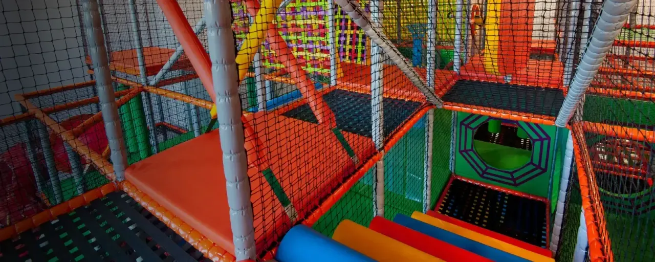 Brightly coloured indoor play structure with netting, platforms and climbing features for children.