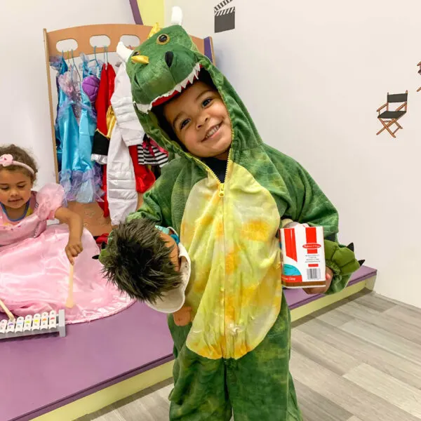 Young child in dinosaur costume holding a toy, smiling at camera in play area with costume rack behind.