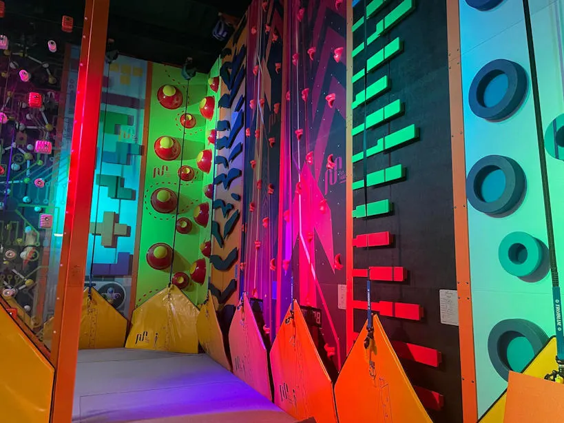 Colourful arcade climbing walls and game panels illuminated with neon lighting inside an indoor play venue.