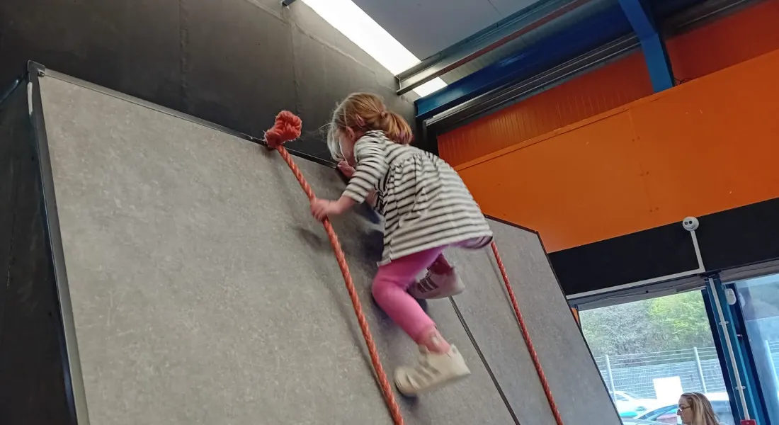 Child climbing a rope wall indoors at an activity centre with coloured ceiling panels.