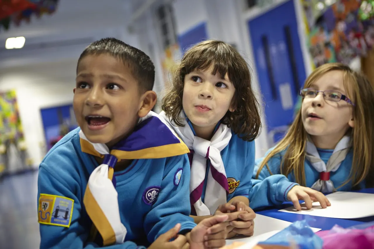 Three young children in blue Scout uniforms sit together at a table, looking upward with engaged expressions during an indoor activity.