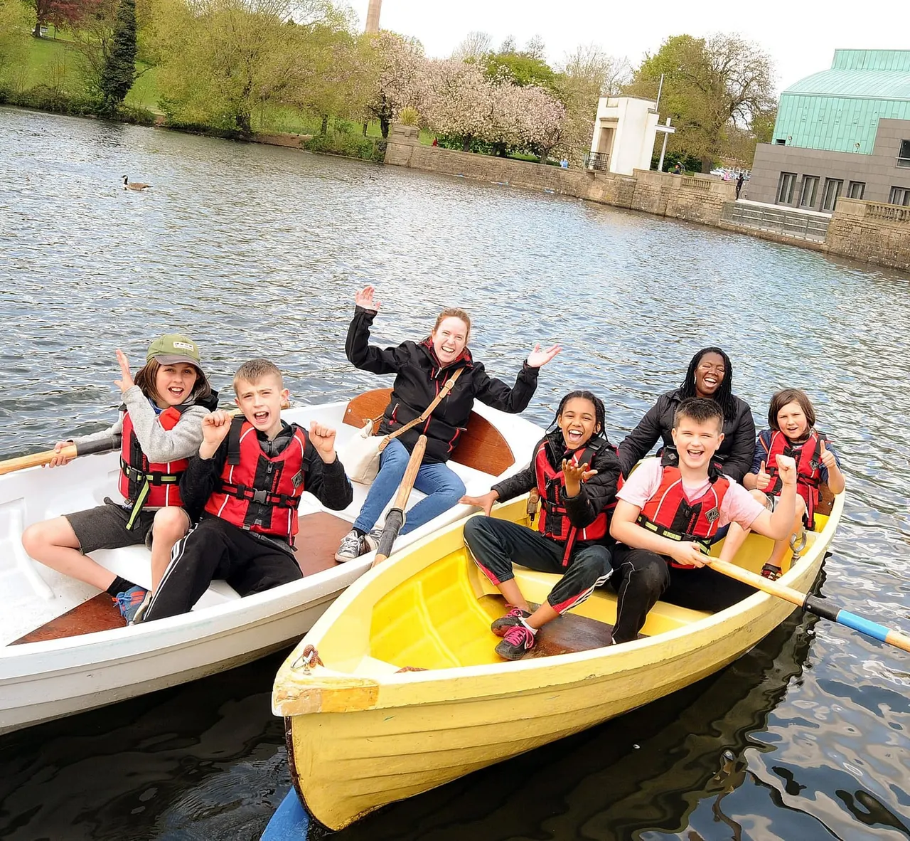 Group of children and young people in life jackets rowing boats on a river or lake, smiling at the camera.