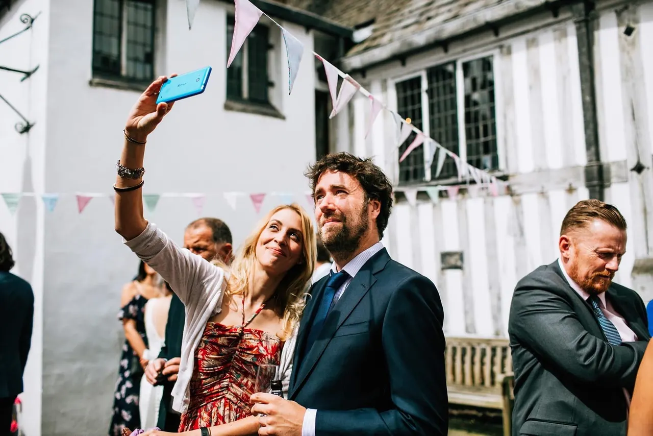 Woman taking a selfie with a man in formal attire at an outdoor event with bunting and a historic building.
