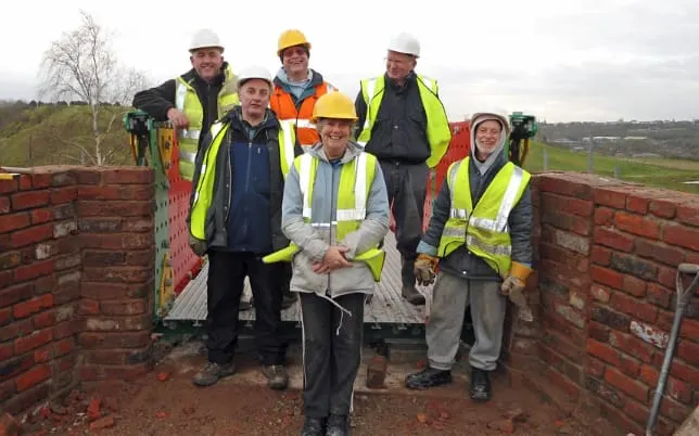 Six workers in high-visibility jackets and hard hats stand together on a brick structure overlooking rural countryside.