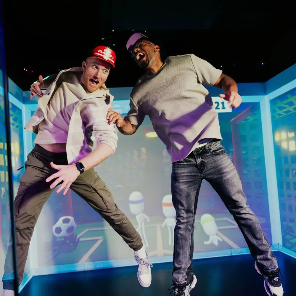Two men in colourful casual wear jump and pose enthusiastically inside an interactive gaming pod with blue-lit virtual sports scenery.