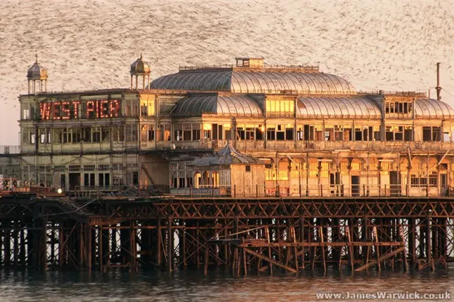 Historic pier buildings with domed roofs lit by golden evening light, viewed from the sea with wooden support structures visible below.
