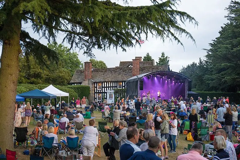 Outdoor concert event with large crowd seated and standing on lawn before illuminated purple stage, historic half-timbered hall visible behind.