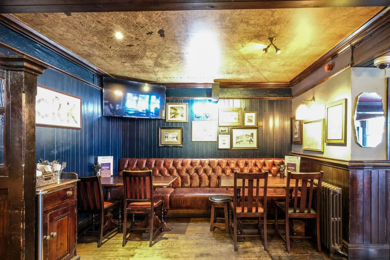 A traditional pub interior with leather banquette seating, wooden chairs, framed pictures on blue-panelled walls, and a textured ceiling lit by warm lights.