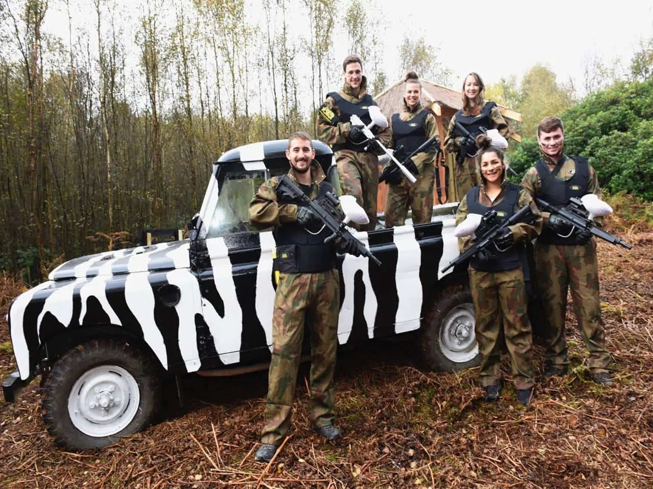 Group of paintball players in camouflage gear posing with airsoft guns beside a zebra-striped vehicle in a woodland setting.