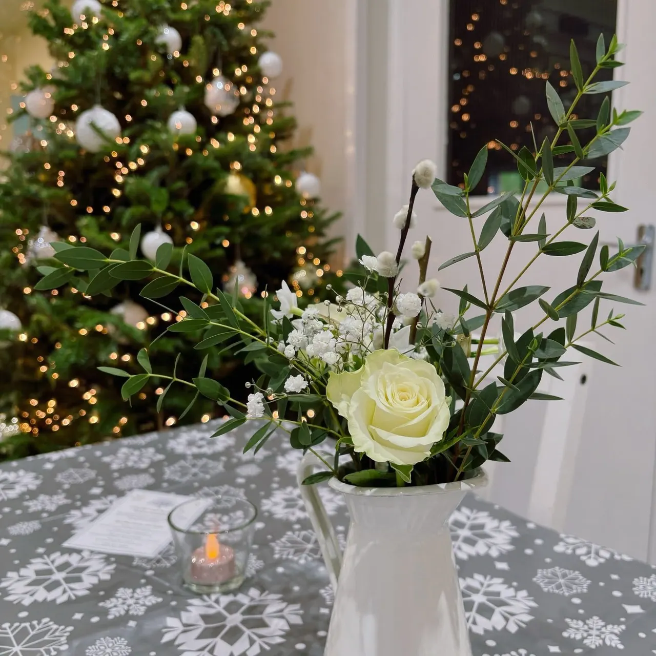 White rose and foliage arrangement in a vase on a festive table, with lit Christmas tree behind.