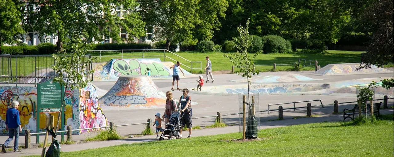 Public park with skate park featuring coloured ramps, visitors including families and young people using the facilities on a sunny day.