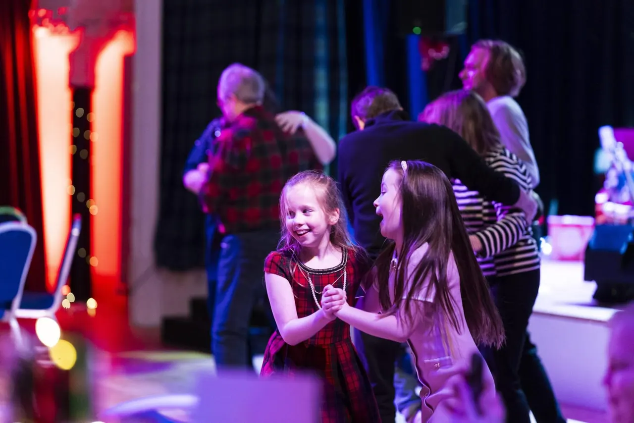 Children dancing together in a colourful lit indoor space with other families in the background.