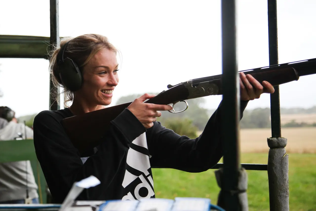 Woman wearing headphones aiming a shotgun at an outdoor clay shooting range with countryside backdrop.