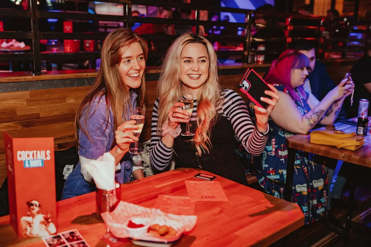 Two young women holding cocktails and smiling at a neon-lit bar counter during a night out.