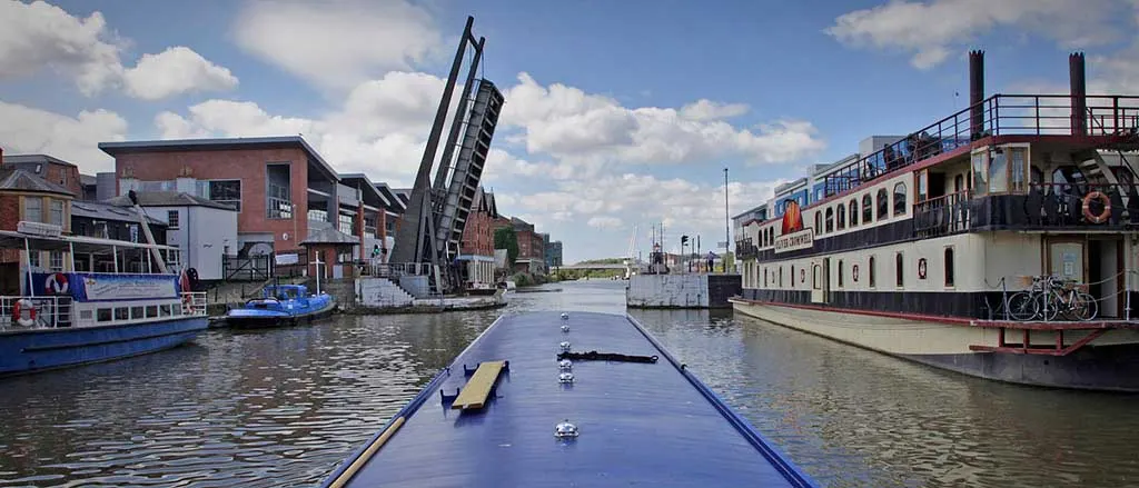 A narrowboat's deck view along a canal basin with moored leisure vessels and a bascule bridge, waterside buildings, and blue sky.
