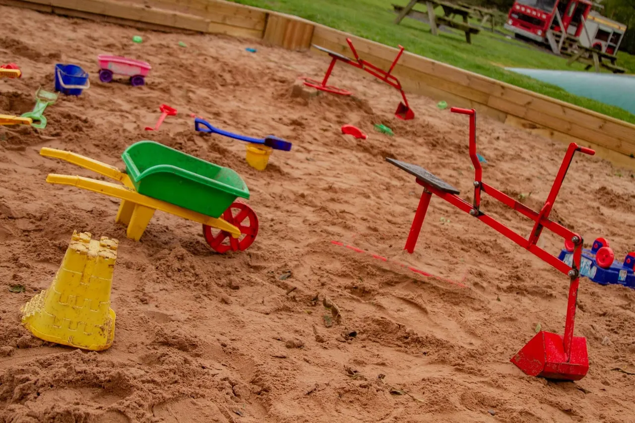 Sandy play area with colourful toy wheelbarrows, diggers and play equipment scattered across the sand.