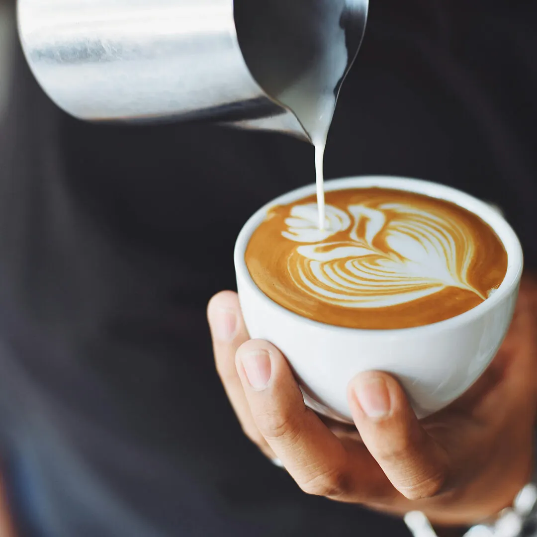 A barista pours steamed milk into a cappuccino with latte art design in a white cup held in someone's hands.